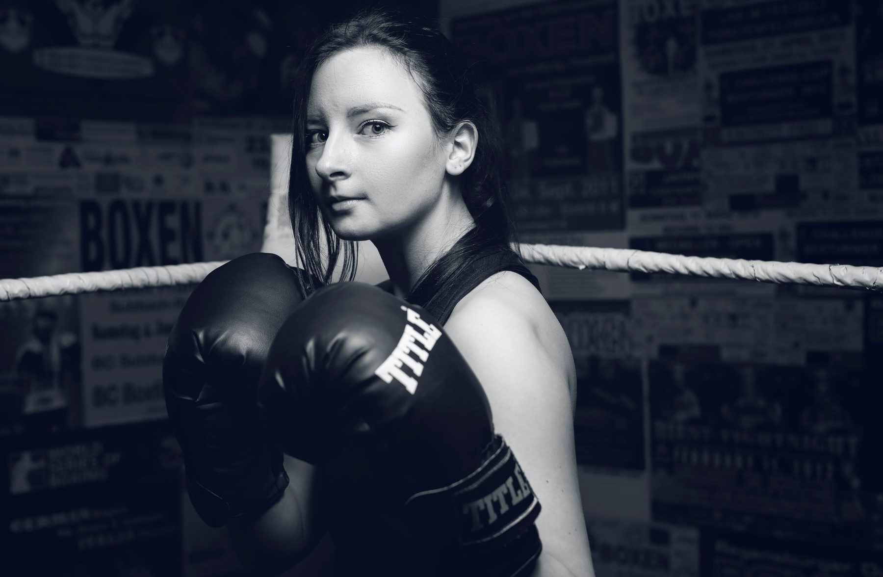 A woman in a vest and black boxing gloves with her hands up, in blue toned light with fight posters behind her