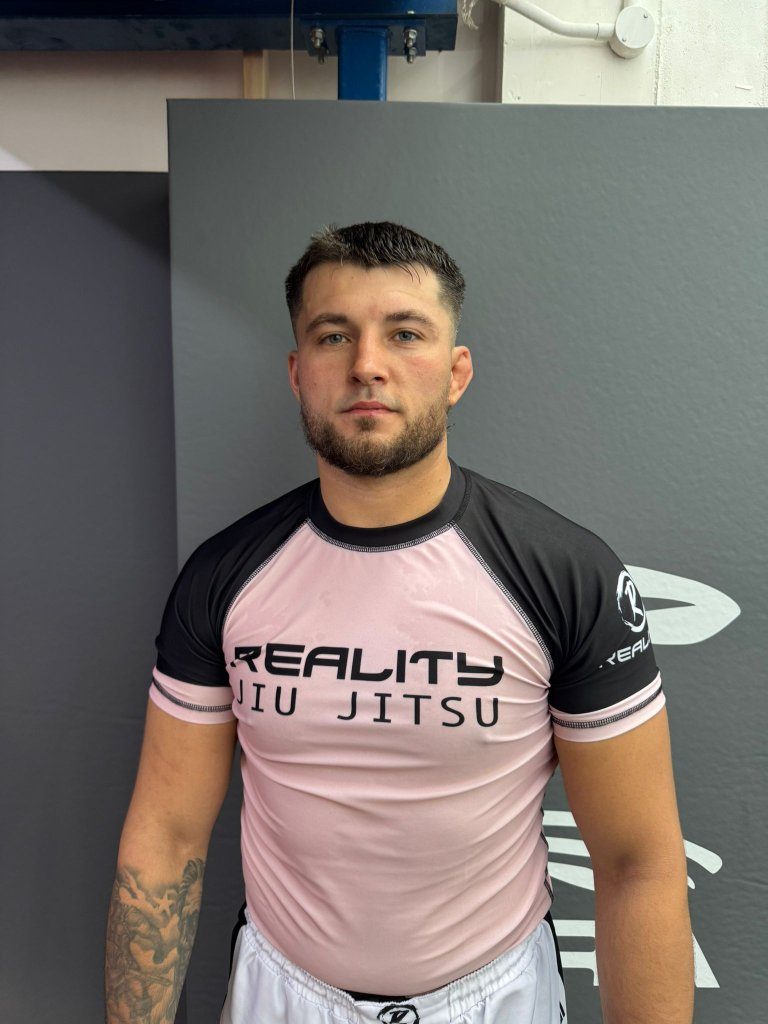 Tommy Collings in a pink Reality Jiu Jitsu rash guard, standing in a gym