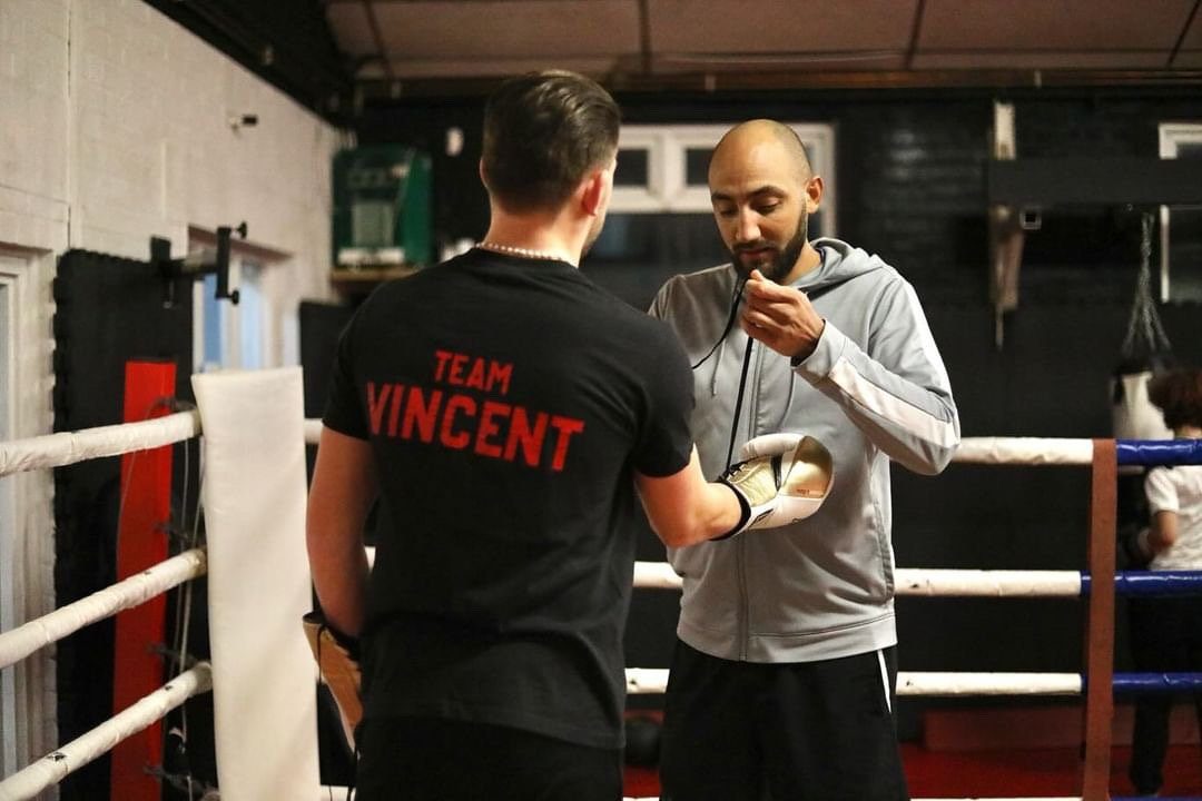 A coach wraps a boxer's hands in the corner of the ring; the boxer's shirt reads Team Vincent