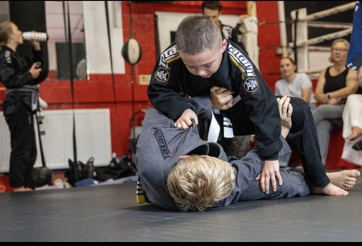 Two children in gis grappling on the mats during a kids' jiu jitsu class, with other students watching