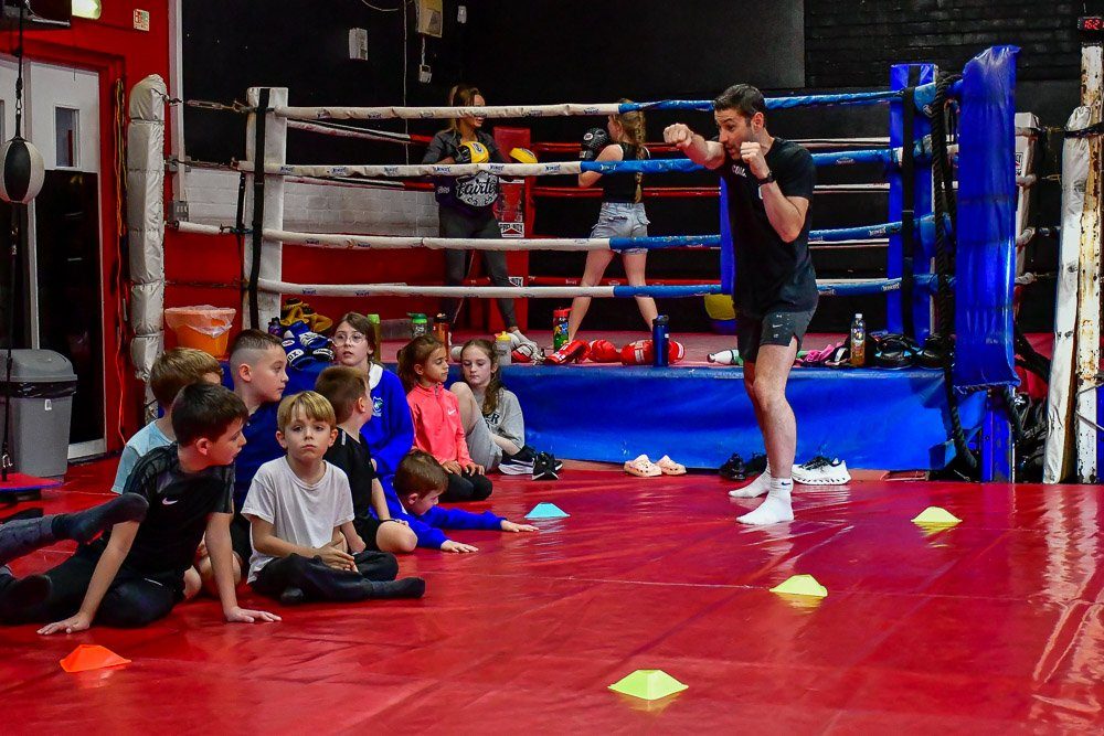 Children seated on the red mats beside the ring while two coaches work in the ring behind them