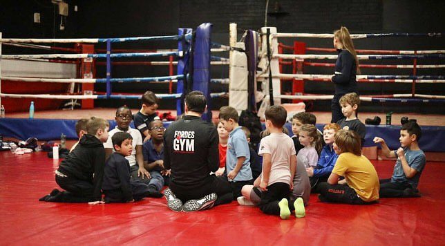 A group of children sitting on the red mats around a coach in a Fordes Gym T-shirt, with the ring behind