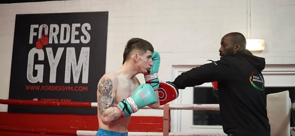 A boxer in teal gloves punches focus mitts held by a coach in front of the Fordes Gym banner