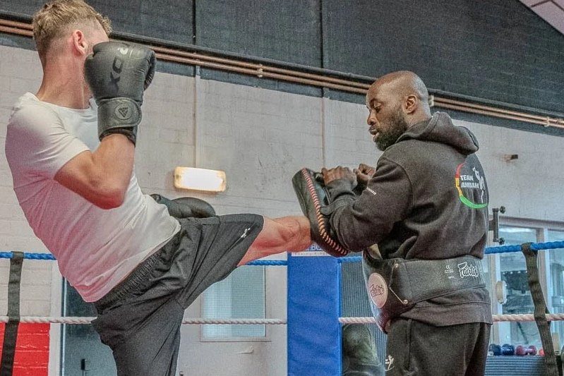 A man in boxing gloves throws a high kick into Thai pads held by a coach in the ring